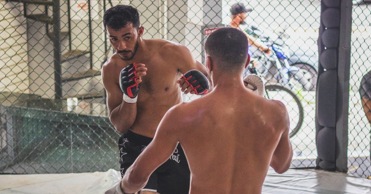 Two fighters spar in a training cage, one wearing open-finger MMA gloves