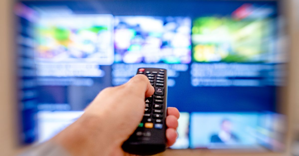 A hand pointing a TV remote at a blurred streaming menu on a television