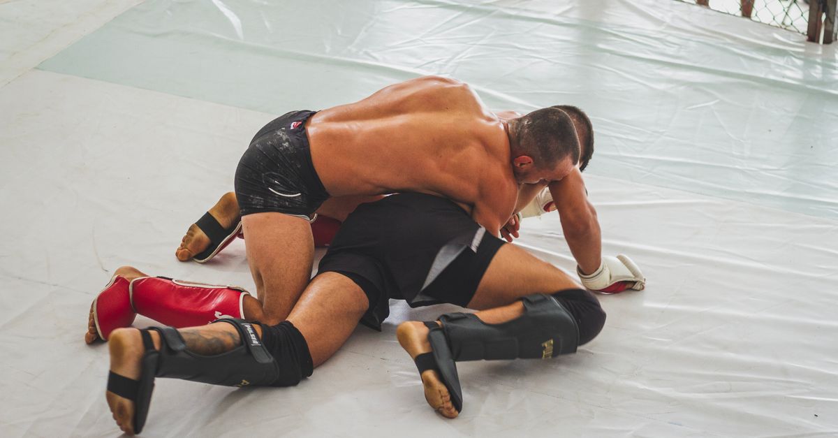 Two MMA fighters in shin guards grappling on the white mat of a training cage