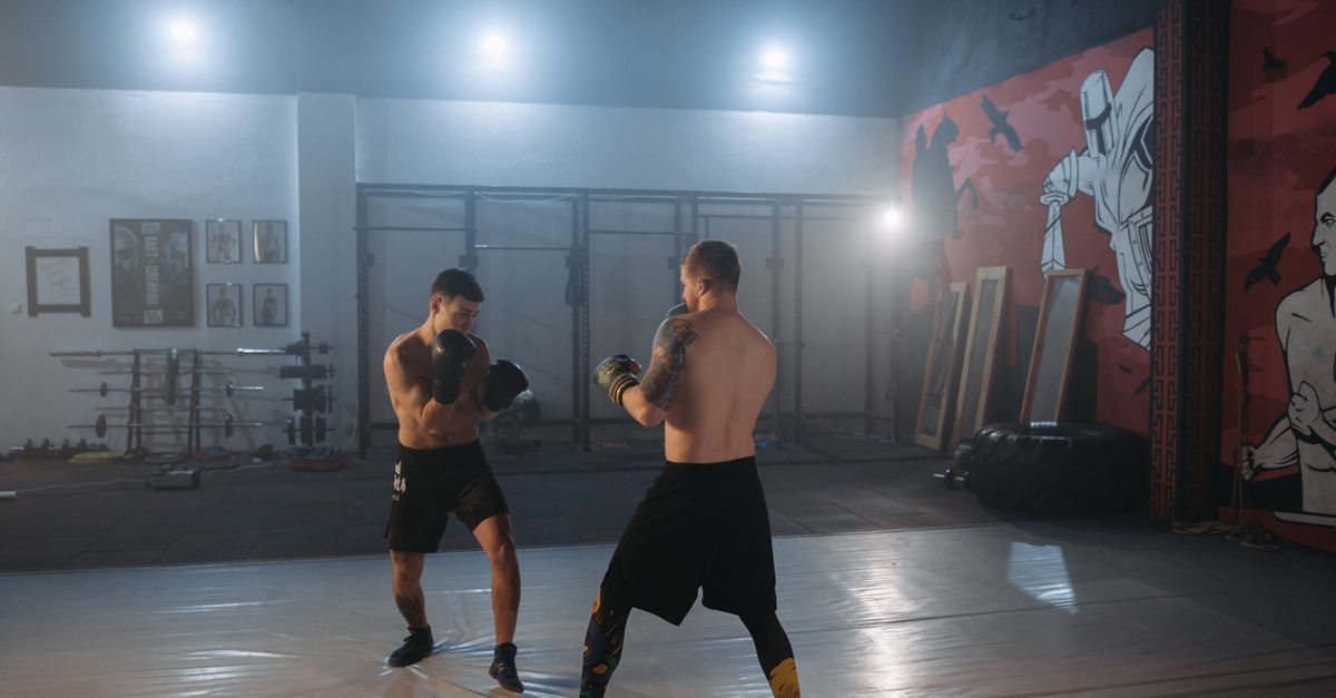 Two shirtless fighters in black shorts sparring on the mat of a large training gym